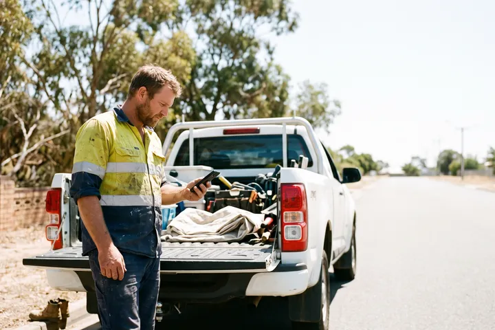 A tradesman checking his phone at the open tailgate of his ute.